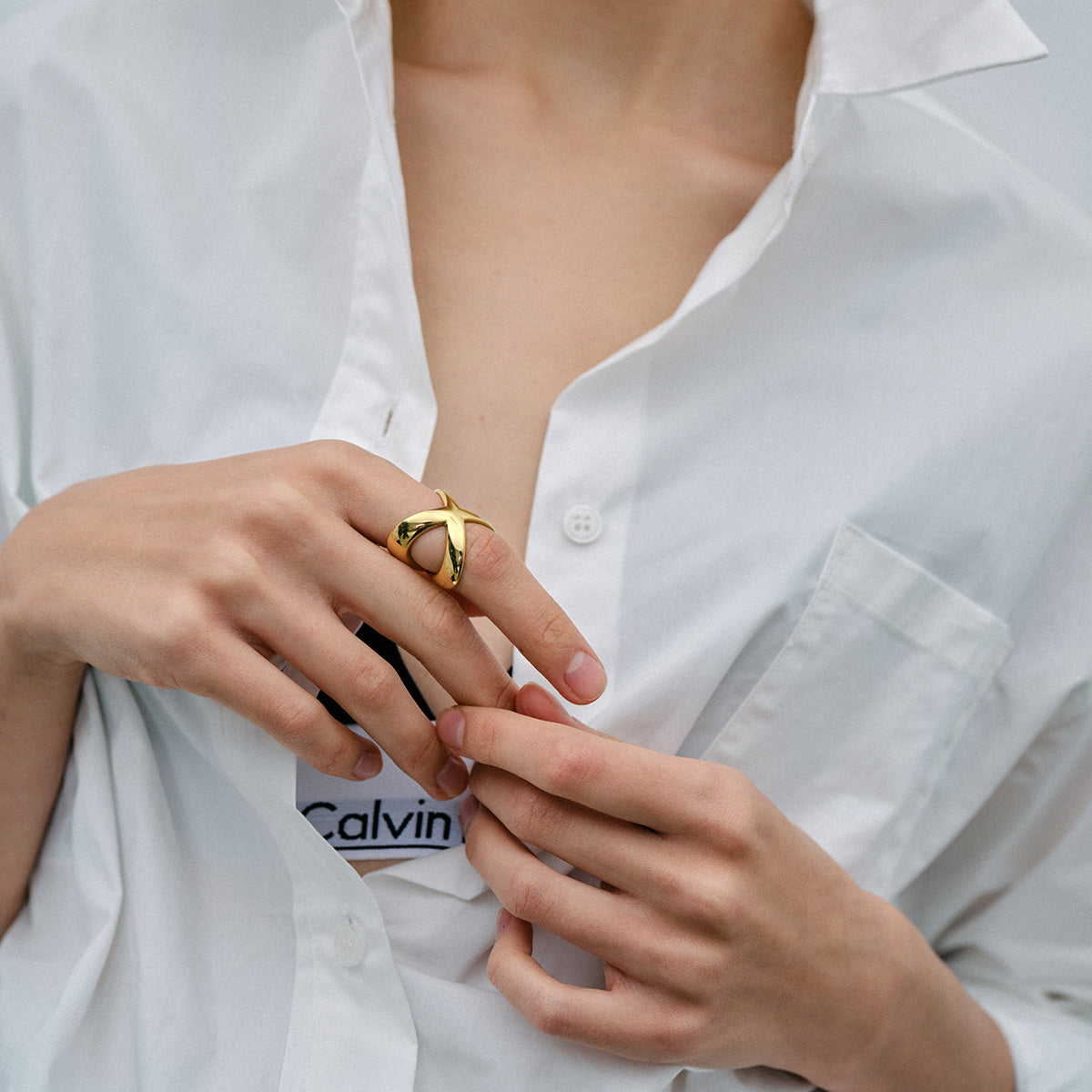 Model showcasing an 18K gold-plated crisscross ring on fingers, wearing a white button-up shirt.
